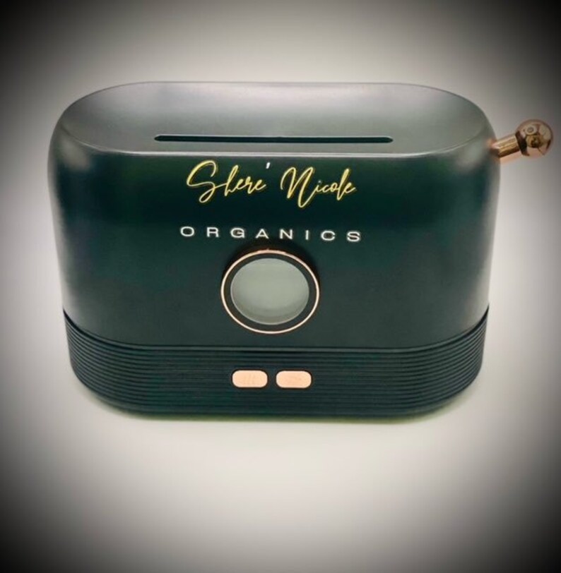 May include: A matte black, oval-shaped electronic device with a top slot, featuring the gold text "Shere' Nicole" and "ORGANICS" printed on the front. It includes a circular display, two rose gold buttons, and a rose gold knob on the right side.