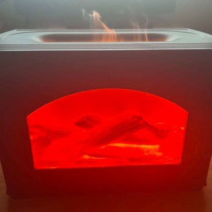 May include: A rectangular black electric fireplace diffuser with a central arched window displaying a bright red glowing light and simulated logs. The top surface features a recessed opening emitting a visible plume of white mist or vapor.