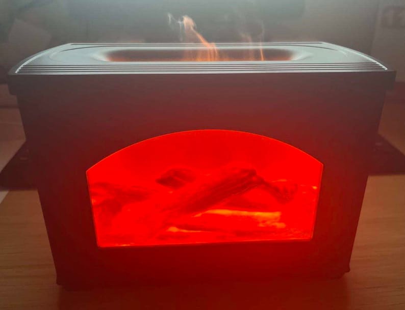 May include: A rectangular black electric fireplace diffuser with a central arched window displaying a bright red glowing light and simulated logs. The top surface features a recessed opening emitting a visible plume of white mist or vapor.