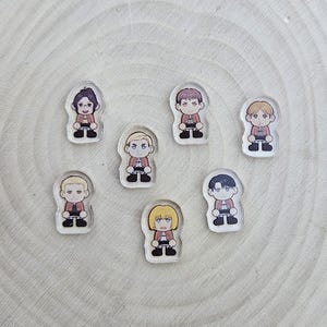 May include: A collection of seven clear acrylic charms featuring cartoon illustrations of characters. Each charm depicts a character in a red jacket, black pants, and black shoes. The characters have different hairstyles and facial expressions.