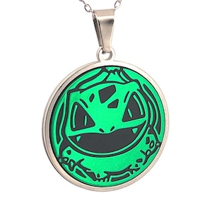 May include: A silver necklace with a circular pendant. The pendant has a green and black design of a smiling face. The necklace is a piece of jewellery. The pendant is approximately 3 cm in diameter.