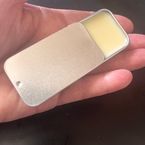 Puede incluir: Una pequeña lata de plata con una tapa con bisagras. La lata contiene un bálsamo amarillo pálido.