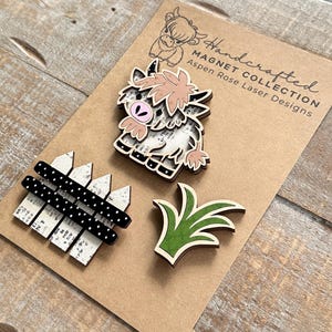 May include: Handcrafted wooden magnets featuring a Highland cow, a fence, and grass. The cow magnet has a pink and beige design. The fence is white with black polka dots. The grass is green with a beige outline. The magnets are on a brown card.