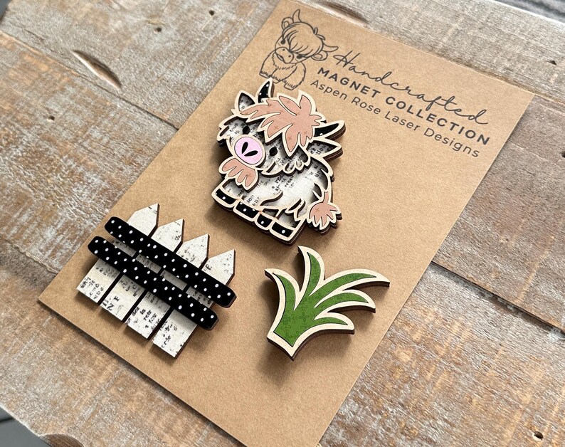 May include: Handcrafted wooden magnets featuring a Highland cow, a fence, and grass. The cow magnet has a pink and beige design. The fence is white with black polka dots. The grass is green with a beige outline. The magnets are on a brown card.