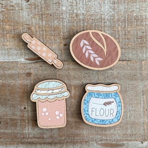 Puede incluir: Recortes de madera con tem&aacute;tica de reposter&iacute;a, que incluyen un rodillo con lunares rosas, una hogaza de pan, un tarro de mermelada y un tarro de harina con la palabra "FLOUR". Los art&iacute;culos son en tonos marr&oacute;n, rosa y azul.