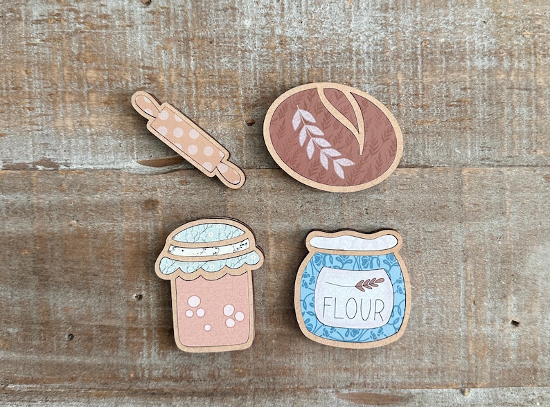Puede incluir: Recortes de madera con tem&aacute;tica de reposter&iacute;a, que incluyen un rodillo con lunares rosas, una hogaza de pan, un tarro de mermelada y un tarro de harina con la palabra "FLOUR". Los art&iacute;culos son en tonos marr&oacute;n, rosa y azul.