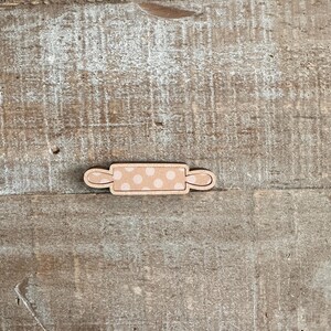 Puede incluir: Un peque&ntilde;o rodillo de madera con acabado marr&oacute;n claro y un dise&ntilde;o de lunares beige. El rodillo tiene asas curvas en cada extremo. El art&iacute;culo est&aacute; sobre una superficie de madera clara y envejecida.