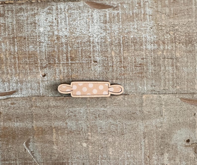 Puede incluir: Un peque&ntilde;o rodillo de madera con acabado marr&oacute;n claro y un dise&ntilde;o de lunares beige. El rodillo tiene asas curvas en cada extremo. El art&iacute;culo est&aacute; sobre una superficie de madera clara y envejecida.