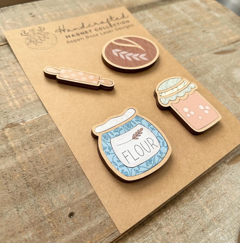 Puede incluir: Un conjunto de imanes de madera hechos a mano con tem&aacute;tica de reposter&iacute;a. Los imanes incluyen un rodillo, un tarro de harina, una taza de caf&eacute; y un tarro con tapa. Los imanes est&aacute;n sobre una tarjeta marr&oacute;n con el texto "Handcrafted Magnet Collection Aspen Rose Laser Designs."
