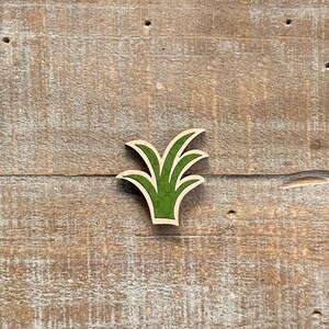 May include: A wooden cutout of a stylized green plant with a light brown border. The plant has multiple pointed leaves and is set against a weathered, light brown wooden background.