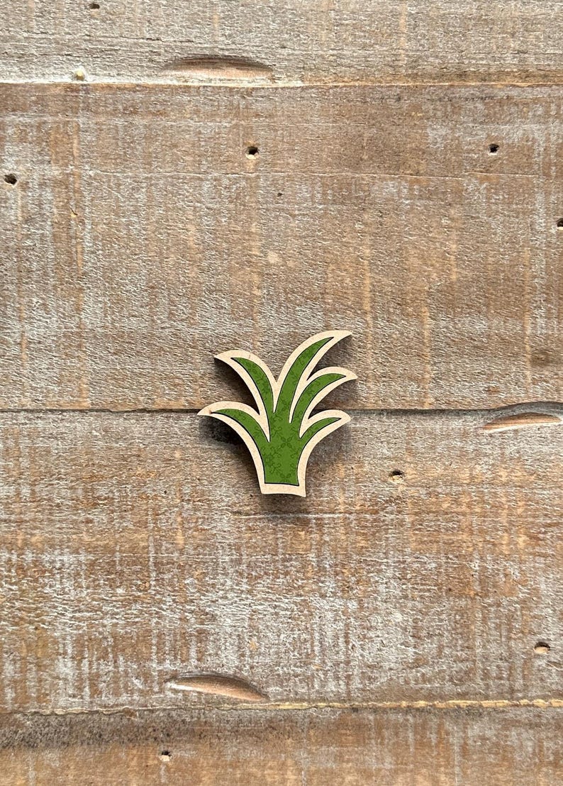 May include: A wooden cutout of a stylized green plant with a light brown border. The plant has multiple pointed leaves and is set against a weathered, light brown wooden background.