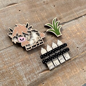 May include: Wooden Highland cow, green grass, and a fence with black polka dots. The cow has a light brown fur, pink nose, and black hooves. The fence has a white picket design.