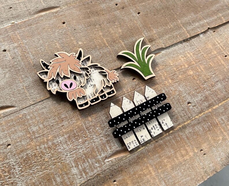 May include: Wooden Highland cow, green grass, and a fence with black polka dots. The cow has a light brown fur, pink nose, and black hooves. The fence has a white picket design.