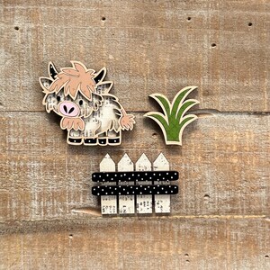 May include: Wooden farm-themed cutouts including a Highland cow, green grass, and a black and white polka dot fence. The cow has a light brown mane and a pink nose. The fence has white picket tops.
