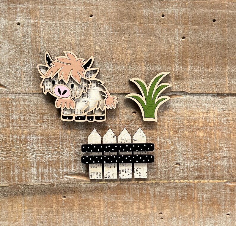May include: Wooden farm-themed cutouts including a Highland cow, green grass, and a black and white polka dot fence. The cow has a light brown mane and a pink nose. The fence has white picket tops.