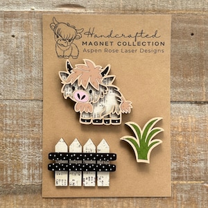 May include: Handcrafted magnet collection featuring a Highland cow, a fence, and grass. The cow has a pink nose and light brown fur. The fence is white with black polka dot accents. The text "Handcrafted Magnet Collection Aspen Rose Laser Designs" is visible.