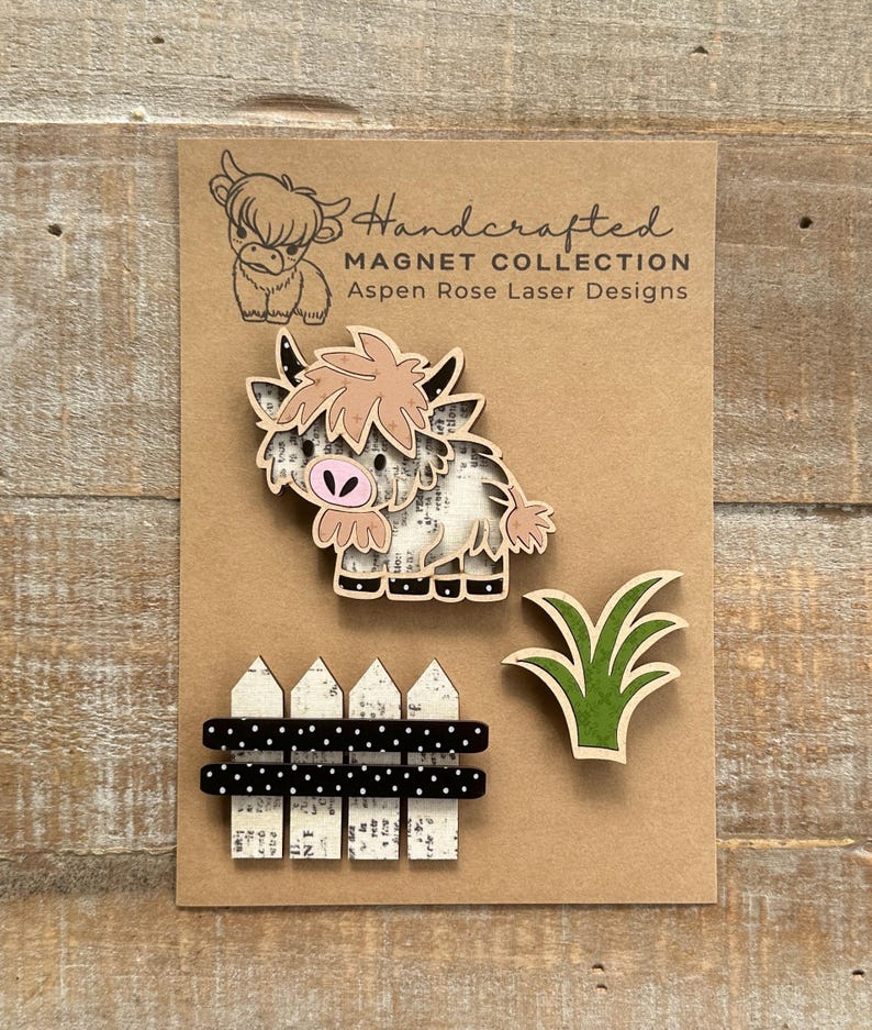 May include: Handcrafted magnet collection featuring a Highland cow, a fence, and grass. The cow has a pink nose and light brown fur. The fence is white with black polka dot accents. The text "Handcrafted Magnet Collection Aspen Rose Laser Designs" is visible.