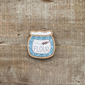 Puede incluir: Una pegatina de madera con forma de tarro, con la palabra "FLOUR" en blanco. El tarro tiene un dise&ntilde;o floral azul y un borde marr&oacute;n. Una peque&ntilde;a ramita de trigo est&aacute; encima de la palabra "FLOUR".