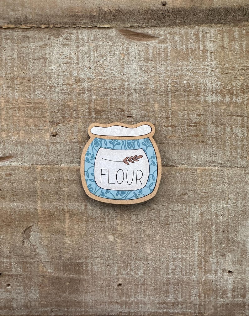 Puede incluir: Una pegatina de madera con forma de tarro, con la palabra "FLOUR" en blanco. El tarro tiene un dise&ntilde;o floral azul y un borde marr&oacute;n. Una peque&ntilde;a ramita de trigo est&aacute; encima de la palabra "FLOUR".
