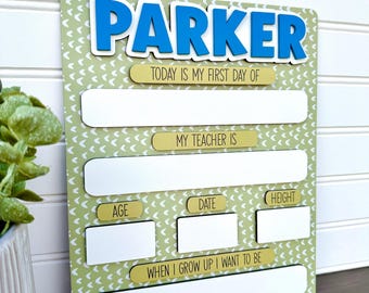 May include: A personalized first day of school sign with the name "PARKER" in blue 3D letters. The sign has spaces to fill in details like teacher's name, age, date, and height. The background is light green with a white pattern.