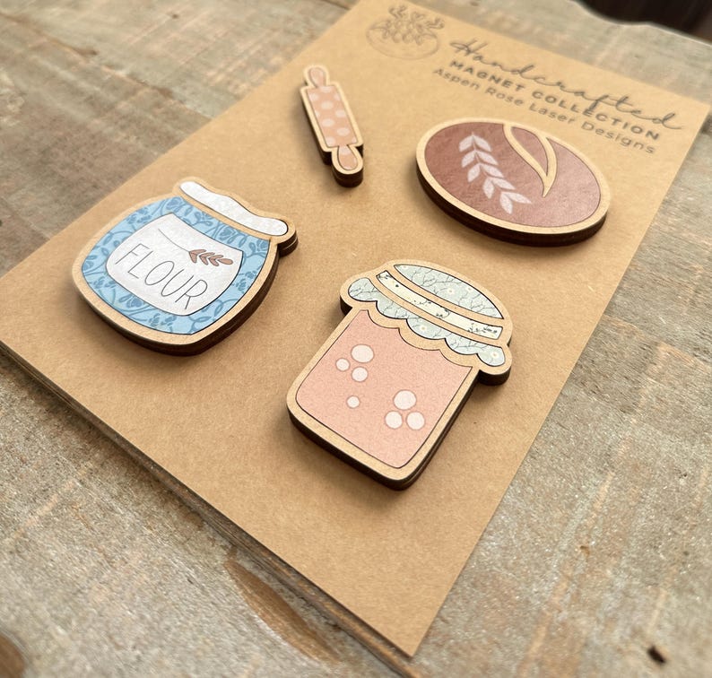 Puede incluir: Imanes de madera hechos a mano con un rodillo, una barra de pan, un tarro de harina y un tarro de mermelada. Los imanes tienen un borde de madera marr&oacute;n claro con colores pastel y dise&ntilde;os florales. El tarro de harina tiene la palabra "FLOUR".
