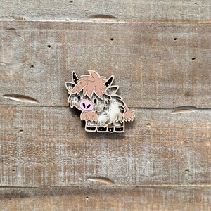 May include: A wooden Highland cow-shaped pin with a light brown and white pattern. The cow has a pink nose and light brown hair. The pin is set against a weathered wood background.