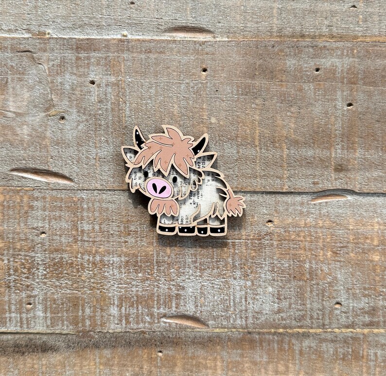May include: A wooden Highland cow-shaped pin with a light brown and white pattern. The cow has a pink nose and light brown hair. The pin is set against a weathered wood background.