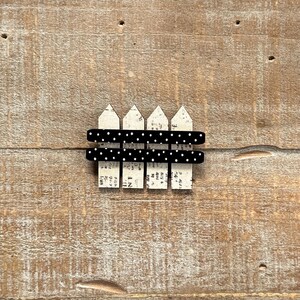 May include: A miniature decorative fence with a distressed white picket design and black polka dot accents. The fence is set against a rustic wooden background, creating a charming, vintage aesthetic.