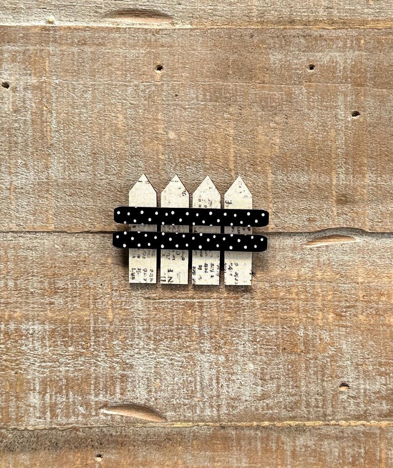 May include: A miniature decorative fence with a distressed white picket design and black polka dot accents. The fence is set against a rustic wooden background, creating a charming, vintage aesthetic.