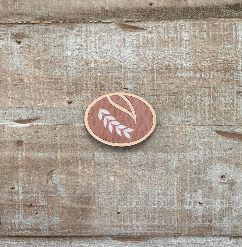 Puede incluir: Un broche ovalado de madera con un dise&ntilde;o de espiga de trigo. El broche presenta un borde marr&oacute;n claro, un fondo estampado y una espiga de trigo blanca. Se encuentra sobre una superficie de madera envejecida.