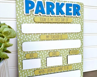 Op de afbeelding: Een rechthoekig bord voor de eerste schooldag met een groen en wit chevronpatroon. De naam PARKER staat in blauwe 3D-letters bovenaan. Het heeft lege witte ruimtes voor het invullen van leeftijd, datum, lengte, leraar en toekomstige beroepswens.