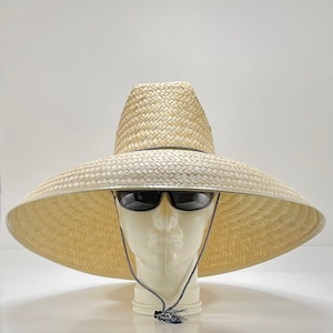 以下が含まれることがあります： 黒いバンドとあご紐が付いた、つば広の麦わら帽子。帽子は白いマネキンの頭に置かれています。