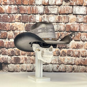 May include: A gray and black cowboy hat with a brown band sits on a white skull mannequin. The skull is wearing sunglasses. The hat is on a white stand against a brick wall background.