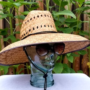 以下が含まれることがあります： 茶色の織り模様と黒いバンドが付いた、つば広の麦わら帽子。帽子は、サングラスをかけたマネキンの頭に載せられています。