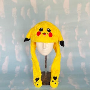 May include: A yellow plush hat with long ear flaps, designed to resemble a popular cartoon character. The hat features black-tipped ears, red cheek circles, and a smiling face. The ear flaps end with black heart-shaped details.