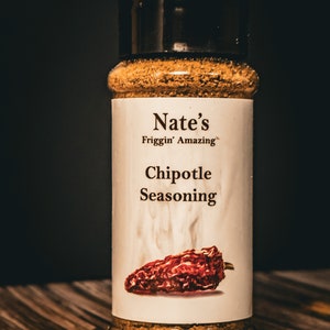 Può includere: Un barattolo di vetro di miscela di spezie marrone con un'etichetta bianca che recita "Nate's Frigg'n Amazing Chipotle Seasoning". L'etichetta presenta un peperoncino rosso con fumo che sale da esso.