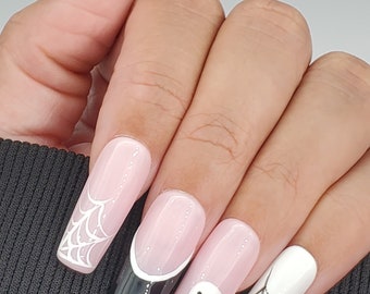 May include: Close-up of a hand with long, pink, almond-shaped nails decorated with Halloween-themed designs. The nails feature white spiderwebs, a black and white spiderweb, a black and white ghost, and a black and white spiderweb.