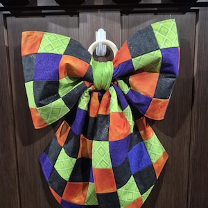 May include: A large fabric bow featuring a patchwork pattern of squares in black, bright orange, purple, and lime green. The fabric squares have varied textures, including solid colors and subtle geometric prints. The bow is gathered at the center.