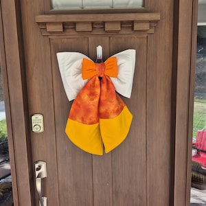 May include: A decorative fabric bow featuring a white top section, a central orange knot, and two long, hanging tails. The tails have a mottled orange and red upper portion and solid yellow lower ends, creating a candy corn-inspired color scheme.