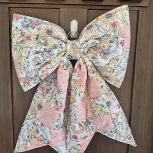 May include: A large decorative bow made of fabric featuring a patchwork design. The pattern consists of alternating sections of white fabric with a colorful floral and paisley print in shades of pink, yellow, and blue, and solid light pink squares.