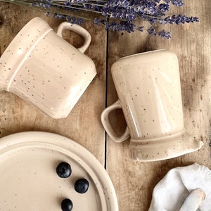 Peut inclure: Trois tasses en céramique beige mouchetées et une assiette en céramique beige mouchetée avec trois myrtilles dessus. Les tasses ont un glaçage beige moucheté et une anse. L'assiette a un glaçage beige moucheté et un bord surélevé. L'arrière-plan est une surface en bois avec un bouquet de lavande.