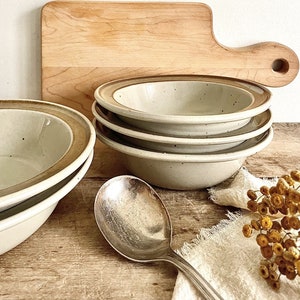 May include: Four beige ceramic bowls with brown rims are stacked, alongside a large silver spoon and a wooden chopping board. Dried yellow flowers and a white cloth are also visible. The bowls are suitable for serving soup or cereal.