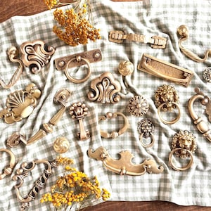 May include: A collection of antique brass hardware, including handles, pulls, and knobs, displayed on a checkered fabric. The hardware features ornate designs, such as lion heads, floral patterns, and ring pulls. Yellow flowers are scattered around the hardware.