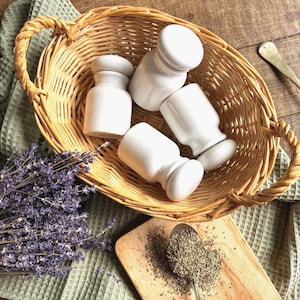 Peut inclure: Un panier en osier tressé contient quatre salières et poivrières en céramique blanche. Une planche à découper en bois avec une cuillère d'herbes et un bouquet de lavande sont également dans l'image. Le panier et la planche sont posés sur un tissu vert.