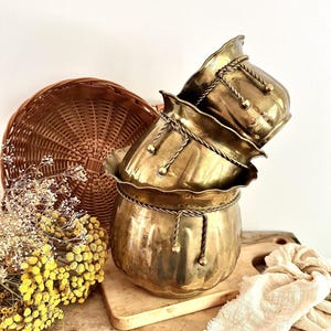 Peut inclure: Une pile de trois cache-pots en laiton avec un bord festonné et une garniture décorative en forme de corde avec des pompons suspendus. Les cache-pots ont une finition métallique polie et un corps arrondi et nervuré en disposition étagée.