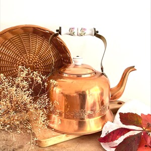 May include: A polished copper tea kettle featuring a wide base, a curved spout, and a metal handle topped with a white ceramic grip decorated with small pink and green floral patterns. A small white ceramic knob sits atop the removable lid.