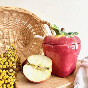 May include: A red ceramic apple-shaped sugar bowl with a lid and a white handle. The bowl is decorated with green leaves and a white flower. A red apple half is on a wooden chopping board next to the bowl.