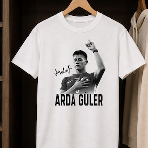 May include: A white short-sleeved crew neck t-shirt featuring a black and white graphic of a soccer player with one hand on their chest and the other raised. The print includes a signature and the text ARDA GÜLER in bold black capital letters.