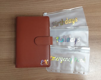 May include: A brown leather planner with a clear plastic pocket on the front. The pocket has three clear plastic envelopes with the words "Birthdays", "Holidays", and "Emergency Fund" written on them.