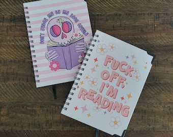 May include: Two spiral bound notebooks with pink and white striped covers. The first notebook has a pink and purple illustration of a skeleton holding a book with the text "Don't judge me by my book covers". The second notebook has a white background with pink flowers and the text "Fuck off, I'm reading".